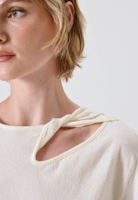 Top color crema con vestibilità rilassata, caratterizzato da un distintivo dettaglio annodato al collo e un tessuto morbido e testurizzato.