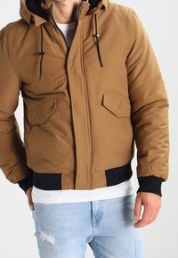 Brown puffer jacket with a hood, featuring black accents, two front pockets, zipper closure, and ribbed cuffs. Worn over a white shirt.