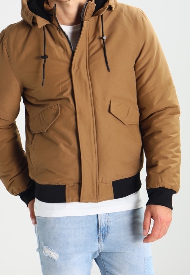 Brown puffer jacket with a hood, featuring black accents, two front pockets, zipper closure, and ribbed cuffs. Worn over a white shirt.