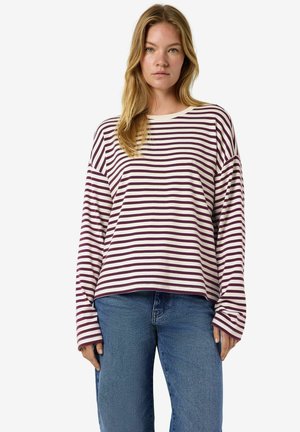 Jeune femme aux cheveux longs portant une chemise blanche ample à manches longues avec des rayures violettes et un jean bleu, debout devant un fond uni.