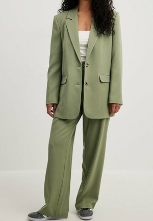 Mujer con americana oversize verde claro y pantalones anchos a juego con zapatillas grises, de pie frente a un fondo claro y liso.