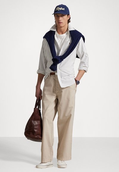 White striped shirt layered with a navy sweater, cream wide-leg pants, white sneakers, and a brown leather bag. Navy cap with logo.