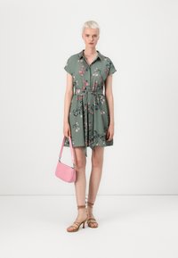 Vestito verde a maniche corte con motivo floreale, cintato in vita. La modella tiene una borsa rosa, indossando sandali con tacco alto e fasce marroni.