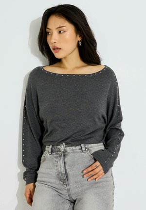 Mujer con cabello largo y oscuro lleva un suéter gris oscuro con pequeños tachones en las mangas y el escote, combinado con vaqueros de tiro alto en gris claro.