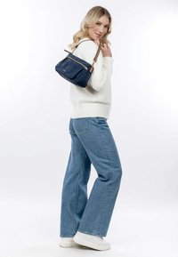 Marineblaue Lederhandtasche mit einem goldenen Reißverschluss, ausgestattet mit einer Fronttasche und braunen Lederelementen. Das Model trägt einen weißen Pullover, Jeans und weiße Sneakers.