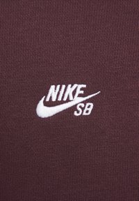 Tecido castanho com textura canelada, apresentando um logótipo Nike SB branco bordado no canto inferior direito.