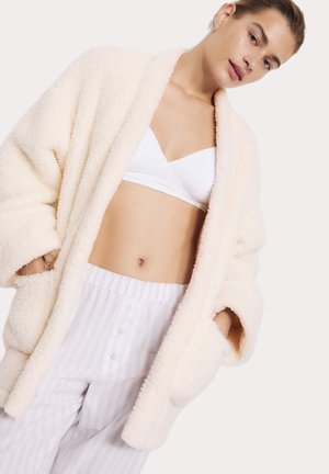 Ung kvinde iført en hvid bralette, hvide pyjamasbukser med striber og en cremefarvet, blød åben badekåbe med hænderne i lommerne.