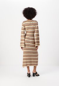 Robe longue et ajustée avec un motif en zigzag dans les couleurs orange, marron et crème. Manches évasées et longueur mi-mollet. Portée avec des sandales à talons carrés.