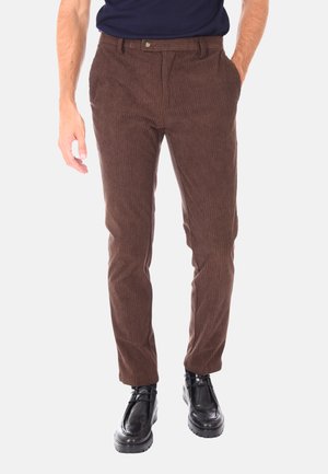 Uomo che indossa pantaloni in velluto a coste marroni, scarpe nere con lacci e una camicia blu navy, in piedi con una mano in tasca.