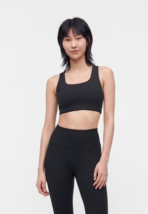 Ung kvinde med sort hår har en sort sports-bh og sorte højtaljede leggings på, stående mod en ensfarvet lys baggrund.