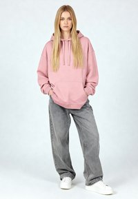 Lichtroze hoodie met voorzak en aantrekkoorden, gecombineerd met losse grijze jeans en witte sneakers, tegen een neutrale achtergrond.