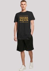 Melna T-krekls ar zelta burtiem "HAKUNA MATATA", virs un zem kura ir ģeometriski raksti; pārī ar melniem šortiem un baltiem sporta apaviem.