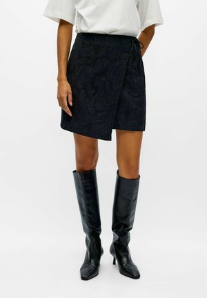 Modèle portant un haut blanc, un short noir texturé à coupe portefeuille et des bottes en cuir noir montant jusqu’aux genoux, sur un fond uni.