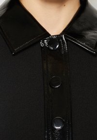 Manteau noir avec col en cuir verni brillant et patte de boutonnage, avec trois grands boutons ronds noirs, porté par une personne.