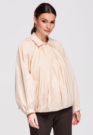 Femme aux cheveux foncés en queue de cheval basse portant un chemisier ample beige clair à manches longues bouffantes et un pantalon foncé sur fond uni.