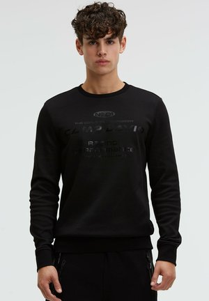 Young man with curly hair wearing a black sweatshirt displaying subtle dark text "Camp David Racing Performance" on front, standing neutral on gray background.