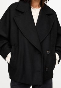 Sort oversize dobbeltradet frakke med store revers, tekstureret stof, to forlommer og knapdetaljer. Bæres over en cremefarvet sweater.