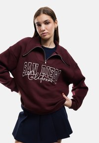 Felpa borgogna con collo alto, design a mezza zip e testo ricamato bianco "SAN DIEGO California." Indossata sopra una gonna blu.
