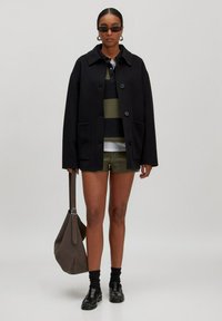 Frau trägt schwarze Jacke, gestreiftes Poloshirt, olivfarbene Shorts, schwarze Socken, Loafer und Sonnenbrille, hält eine große braune Tasche vor einem schlichten Hintergrund.