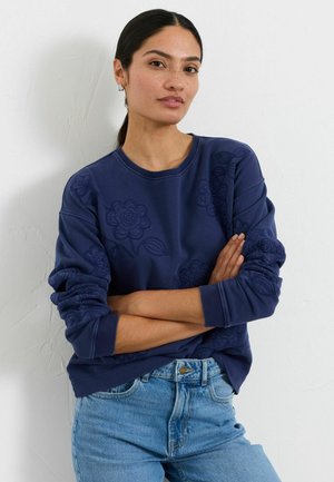 Woman with dark hair in a ponytail wearing a navy blue floral embroidered sweatshirt and light blue jeans, standing with arms crossed.