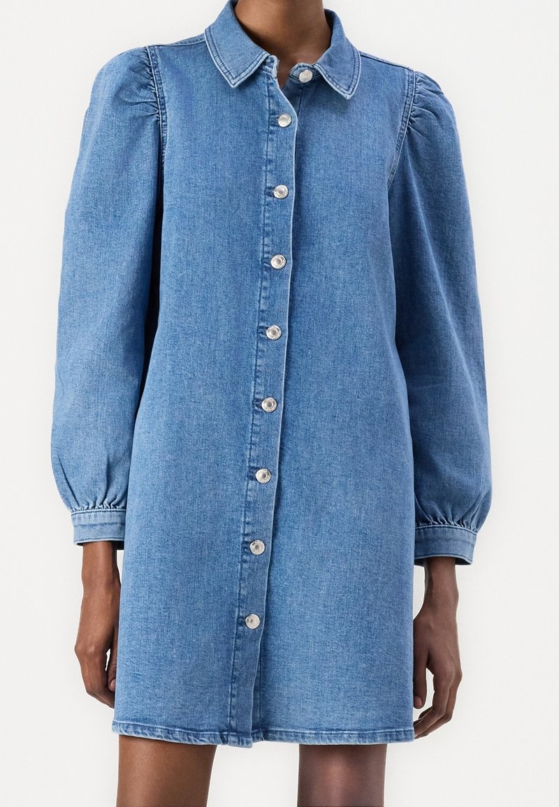 Robe-chemise en denim avec fermeture boutonnée sur le devant, col et manches bouffantes. Couleur bleu clair, texture lisse et longueur mi-cuisse.