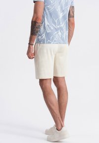 Pantalones cortos de algodón de color claro con un ajuste relajado, combinados con una camisa azul de manga corta con un patrón de hojas. Zapatillas visibles.