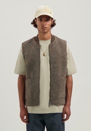 Jonge man in een beige pet, lichtgroene t-shirt, bruine vest met rits en een ketting met twee hangers, staande tegen een effen achtergrond.