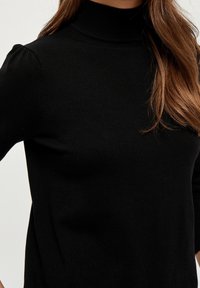 Svart stickad tröja med hög hals, figurnära passform och subtila puffärmar. Slät textur utan synliga mönster eller detaljer.