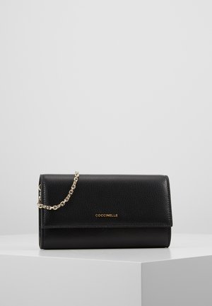 Pochette Coccinelle en cuir noir avec chaîne dorée et logo doré, présentée sur une surface angulaire blanche contre un fond uni.