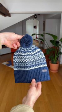 Kék kötött sapka csíkos hópehely mintával, tetején bojttal és bordázott szegéllyel. Puha fonalból készült, textúrával.