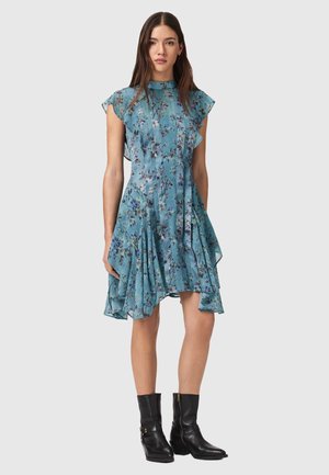 Robe fleurie bleue avec manches courtes évasées, jupe plissée et col montant. Associée à des bottines noires avec des accents dorés.