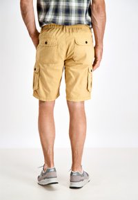 Pantaloni cargo gialli con tasche posteriori, vita elastica e tasche laterali per cargo. Indossati con sneaker grigie e una camicia a scacchi visibile nella parte superiore.