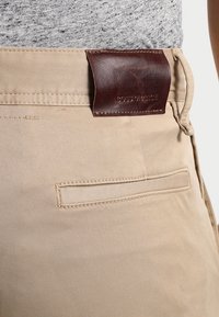 Beige byxor med en slät textur, sömnadsdetaljer och en brun läderetikett i midjan med uppburet tryck.