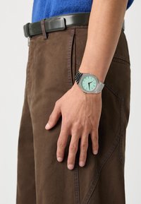 Montre argentée avec un cadran vert menthe et un bracelet à motif noir, portée sur une main contre un pantalon marron, affichant des détails de coutures visibles.