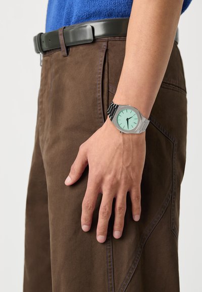Ezüst karóra menta zöld számlappal és fekete mintás szíjjal, egy kézen viselve barna nadrágok mellett, látható varrási részletekkel.