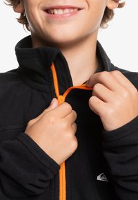 Schwarze Fleecejacke mit einem orangefarbenen Reißverschluss, Hände, die den Kragen anpassen. Hat ein Logo auf der linken Seite und ein hohes Kragen-Design.