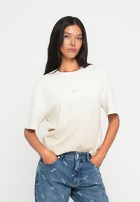 Kermanvärinen t-paita lyhyillä hihoilla ja pyöreällä kaula-aukolla, jossa on huomaamaton vaaleanruskea logo rinnassa.