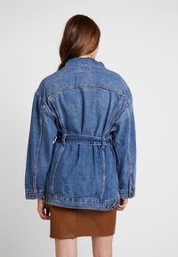 Blå denimjacka med avslappnad passform, långa ärmar, bälte i midjan och dubbla sidofickor. Texturerat tyg med en något blekt look.