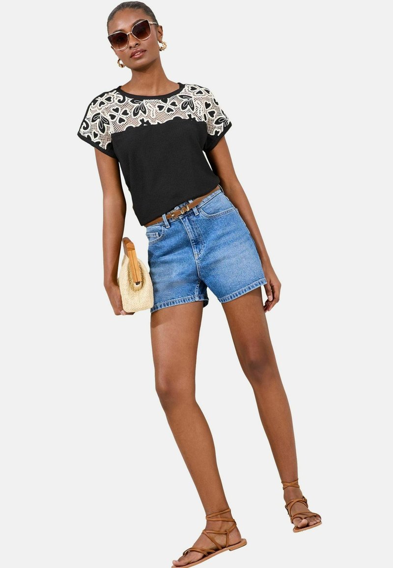 Top negro con detalles de encaje, pantalones cortos de denim y sandalias marrones con tiras. La modelo sostiene un bolso tejido beige, mostrando un atuendo casual de verano.