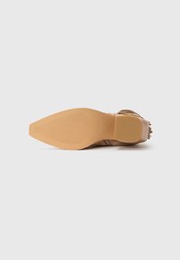 Zapato de tacón de ante marrón con punta afilada, suela estriada y detalles de flecos en el tacón. Textura suave y color natural.