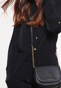 Černá crossbody taška se zlatými detaily, texturovanou kůží a zakřiveným tvarem; obsahuje řetízkový popruh a logo akcent.