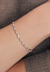 Srebrna bransoletka z naprzemiennie ułożonymi małymi perłami i przezroczystymi kryształkami, delikatny wzór o gładkim wykończeniu, noszona na nadgarstku.
