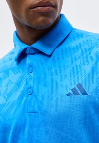 Άνδρας που φοράει μπλε πόλο μπλούζα Adidas με γεωμετρικά σχέδια και πατιλέτα με τρία κουμπιά, κομμένη κάτω από τη μύτη.