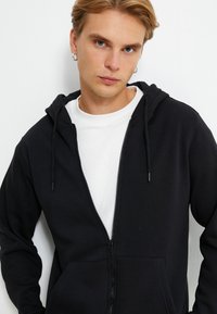 Sudadera con cremallera negra, capucha ajustable con cordón y dos bolsillos laterales. Llevada sobre una camiseta blanca. Textura suave.