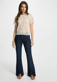 Top in pizzo all'uncinetto color crema, con maniche corte e orlo a scallop. Jeans a zampa d'elefante blu scuro con cuciture verticali e fessure frontali, abbinati a sandali marroni.