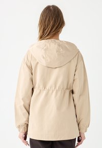Beżowa, lekka kurtka z kapturem, elastycznym pasem i mankietami. Gładki materiał, minimalistyczny design i brak widocznych wzorów.