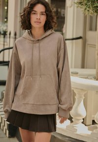 Brązowy zamszowy hoodie z kieszenią kangurką, kapturem na sznurki i ściągaczami, zestawiony z ciemną plisowaną spódnicą.