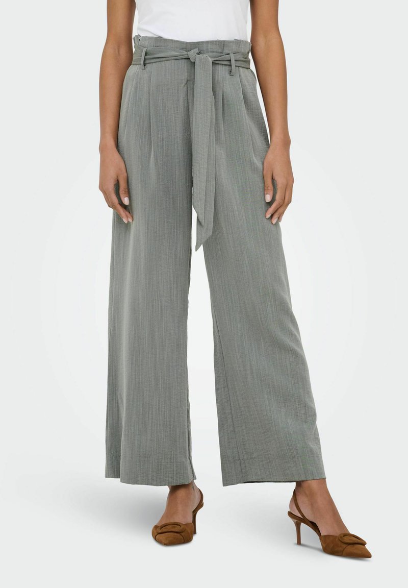 Femme portant un pantalon gris clair taille haute, jambes larges, avec ceinture en tissu, associé à des talons slingback marron et un haut blanc rentré.