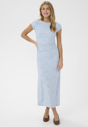 Femme debout, main sur la hanche, portant une robe maxi bleu clair en dentelle à manches courtes et des chaussures tressées marron, devant un fond uni.