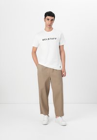 Fehér pamut póló "BELSTAFF" felirattal fekete nyomattal, bézs széles szárú nadrággal és fehér sportcipővel párosítva, egy sima háttér előtt állva.